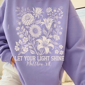 May include: A light purple sweatshirt featuring a floral design in white, including lilies, sunflowers, and other flowers. The text "LET YOUR LIGHT SHINE Matthew 5:16" is printed in white script below the floral arrangement. The brand name "AWEBO" is printed on the left sleeve.
