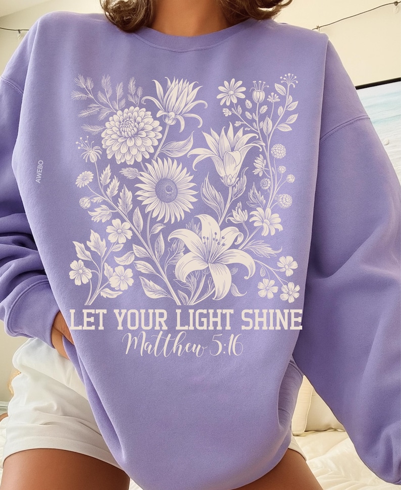 May include: A light purple sweatshirt featuring a floral design in white, including lilies, sunflowers, and other flowers. The text "LET YOUR LIGHT SHINE Matthew 5:16" is printed in white script below the floral arrangement. The brand name "AWEBO" is printed on the left sleeve.