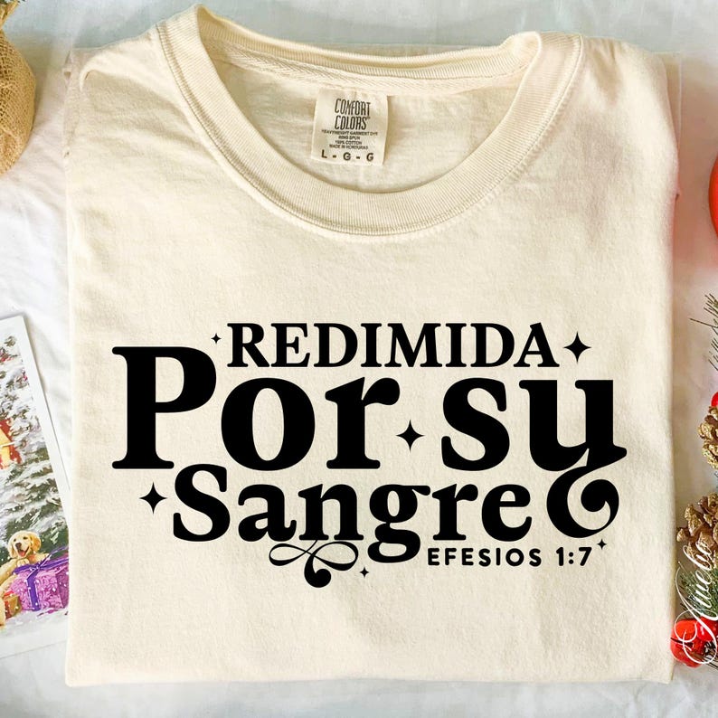 May include: Cream-colored t-shirt with the text "REDIMIDA Por Su Sangre EFESIOS 1:7" in black. The shirt has a crew neck and short sleeves. The Comfort Colors tag is visible on the neckline.