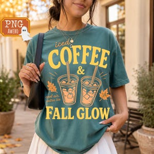 Peut inclure: Un t-shirt à manches courtes bleu sarcelle foncé avec un motif jaune représentant deux verres de café glacé avec des pailles et des glaçons. Le texte indique "iced COFFEE & FALL GLOW" avec "cool air, extra ice" et des feuilles d'automne orange.