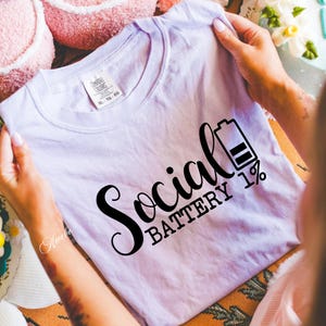 Puede incluir: Una camiseta lavanda con el texto "Social Battery 1%" en letras negras. El texto est&aacute; en una fuente estilizada e incluye un icono de bater&iacute;a con una barra llena.