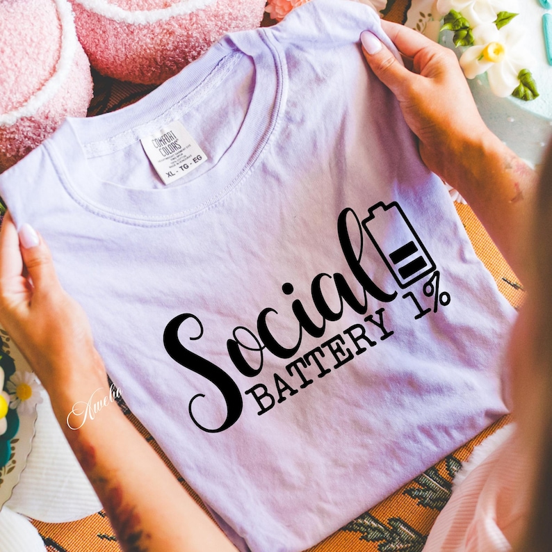 Puede incluir: Una camiseta lavanda con el texto "Social Battery 1%" en letras negras. El texto est&aacute; en una fuente estilizada e incluye un icono de bater&iacute;a con una barra llena.