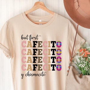 Puede incluir: Una camiseta beige con el texto "but first cafecito y chismecito" en letras negras y marrones. La palabra "cafecito" se repite cuatro veces con diferentes patrones en cada repetici&oacute;n.
