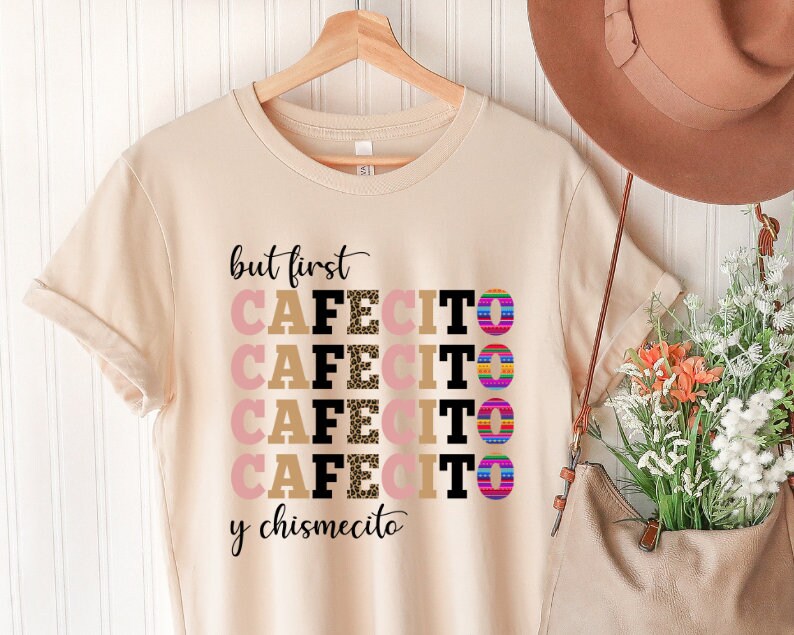 Puede incluir: Una camiseta beige con el texto "but first cafecito y chismecito" en letras negras y marrones. La palabra "cafecito" se repite cuatro veces con diferentes patrones en cada repetici&oacute;n.