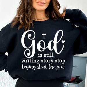 May include: Black sweatshirt with white text that reads "God is still writing story stop trying steal the pen". The text includes a cross and a heart.