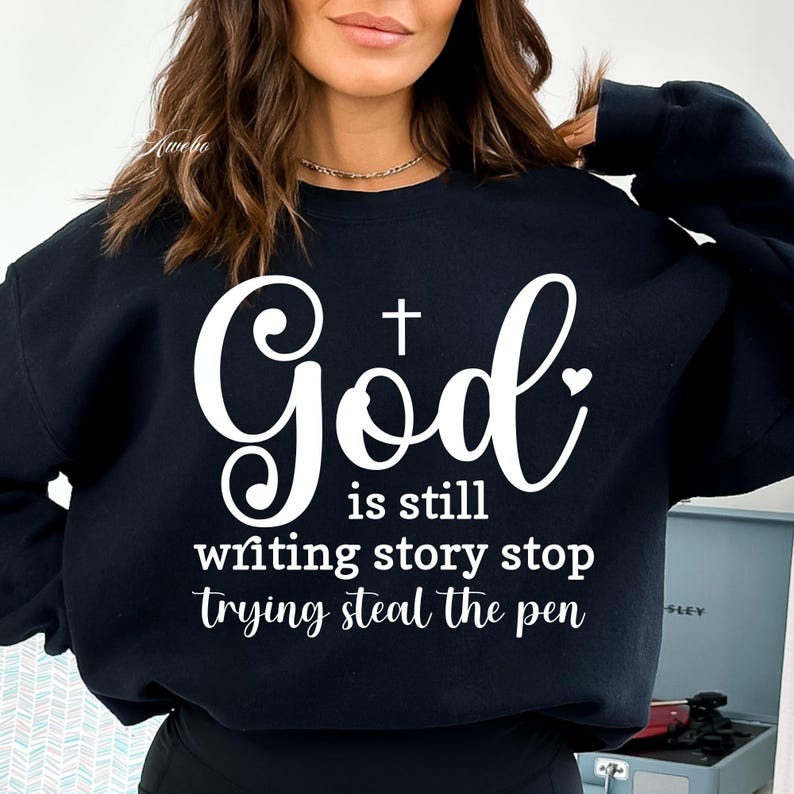 May include: Black sweatshirt with white text that reads "God is still writing story stop trying steal the pen". The text includes a cross and a heart.