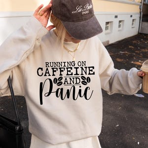 Puede incluir: Una sudadera blanca con el texto "Running on Caffeine and Panic" en letras negras. El texto est&aacute; rodeado por dos s&iacute;mbolos de granos de caf&eacute;.