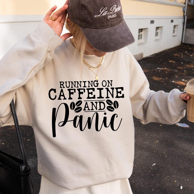 Puede incluir: Una sudadera blanca con el texto "Running on Caffeine and Panic" en letras negras. El texto est&aacute; rodeado por dos s&iacute;mbolos de granos de caf&eacute;.