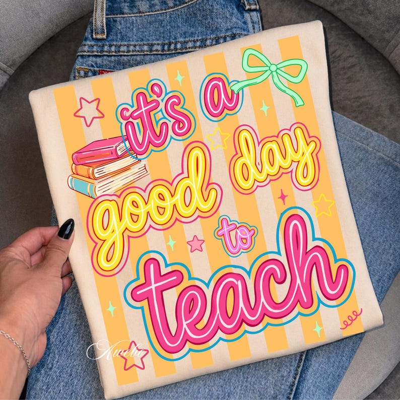 May include: A beige tote bag with the text "It's a good day to teach" in pink and yellow. The bag has a striped background and features illustrations of books, stars, and a bow.