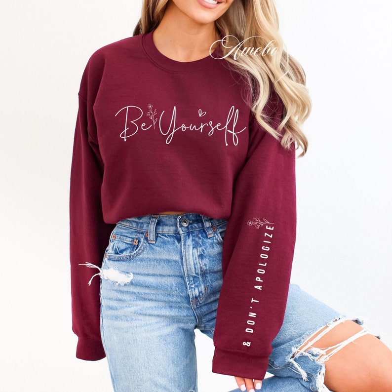 May include: A maroon cropped sweatshirt with the text "Be Yourself" and a small floral design on the front. The sleeves have the text "& Don't Apologize" printed on them.