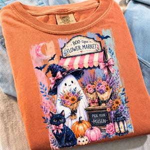 May include: An orange t-shirt featuring a graphic of a ghost wearing a witch hat, a black cat with a pink bow, pumpkins, and a flower market stall. The sign reads Boo-tique Flower Market and Pick Your Poison. The shirt has a Comfort Colors label.