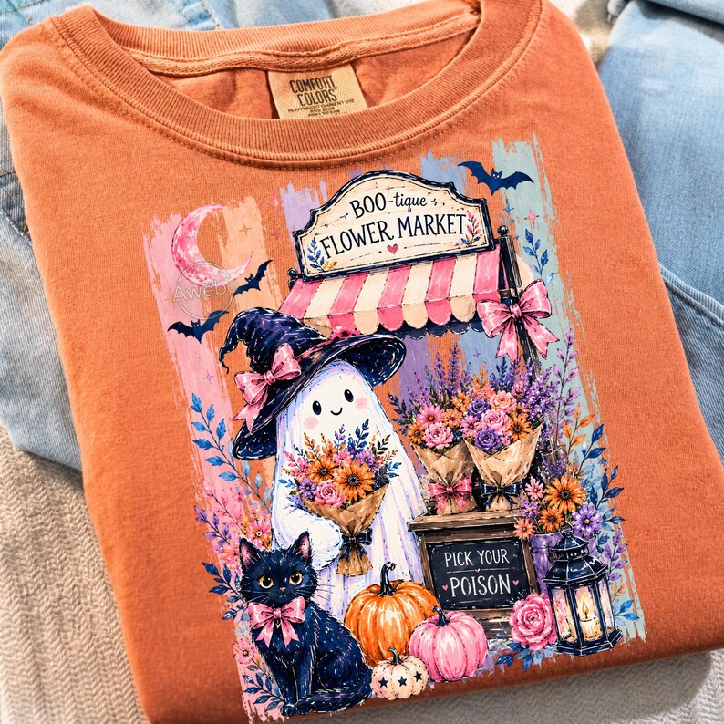 May include: An orange t-shirt featuring a graphic of a ghost wearing a witch hat, a black cat with a pink bow, pumpkins, and a flower market stall. The sign reads Boo-tique Flower Market and Pick Your Poison. The shirt has a Comfort Colors label.