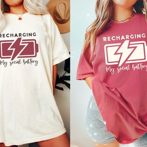 May include: Two women wearing oversized t-shirts with the text "RECHARGING My social battery" and a graphic of a battery with a lightning bolt. The t-shirt on the left is cream colored with maroon text and graphic. The t-shirt on the right is a deep red color with white text and graphic.