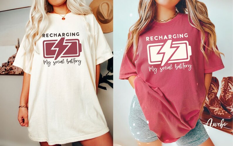 May include: Two women wearing oversized t-shirts with the text "RECHARGING My social battery" and a graphic of a battery with a lightning bolt. The t-shirt on the left is cream colored with maroon text and graphic. The t-shirt on the right is a deep red color with white text and graphic.