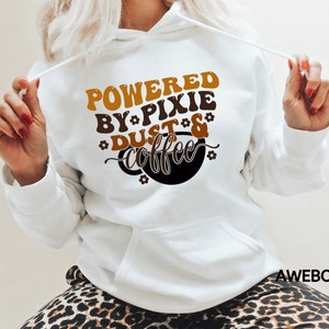 Może przedstawiać: Biała bluza z kapturem z brązowym i pomarańczowym nadrukiem, na kt&oacute;rym widnieje napis "Powered by Pixie Dust & Coffee".