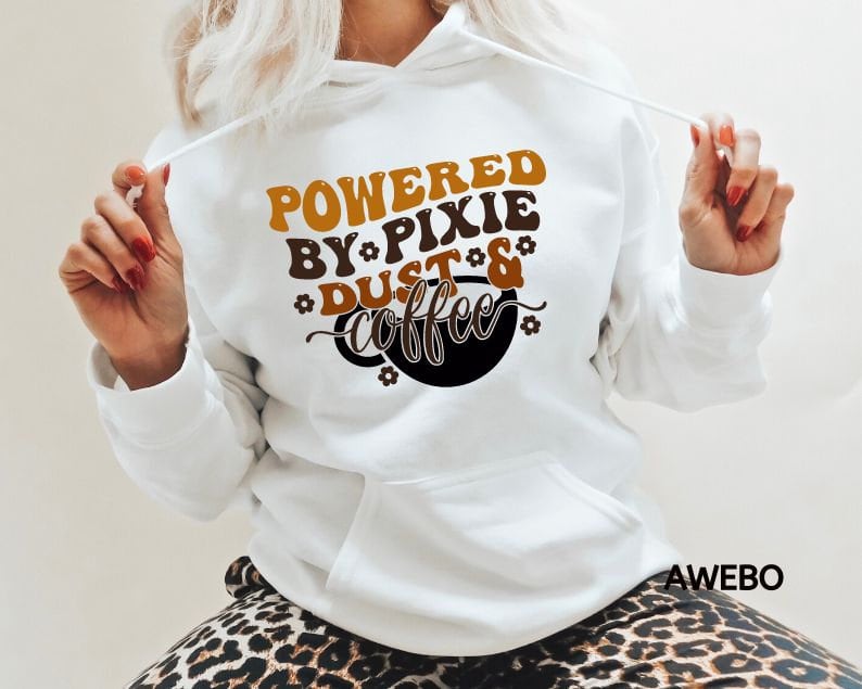 Może przedstawiać: Biała bluza z kapturem z brązowym i pomarańczowym nadrukiem, na kt&oacute;rym widnieje napis "Powered by Pixie Dust & Coffee".