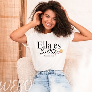 Puede incluir: Una camiseta blanca con el texto "Ella es fuerte" y "Proverbios 31:25" en letras negras. La camiseta presenta un dise&ntilde;o floral con acentos rosas, amarillos y naranjas.