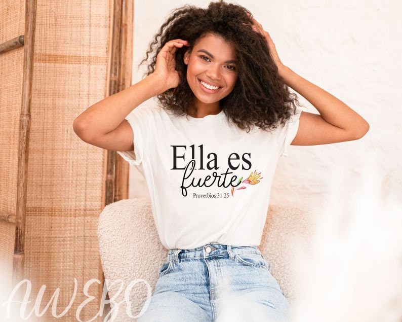 Puede incluir: Una camiseta blanca con el texto "Ella es fuerte" y "Proverbios 31:25" en letras negras. La camiseta presenta un dise&ntilde;o floral con acentos rosas, amarillos y naranjas.