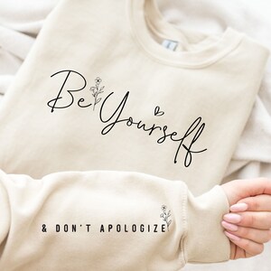 May include: A beige sweatshirt with the text "Be Yourself & Don't Apologize" printed on it. The text is in black cursive font with a small floral design next to the word "Be".