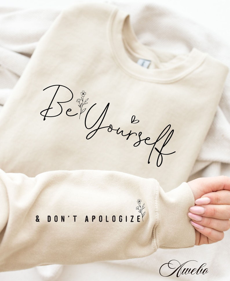 May include: A beige sweatshirt with the text "Be Yourself & Don't Apologize" printed on it. The text is in black cursive font with a small floral design next to the word "Be".