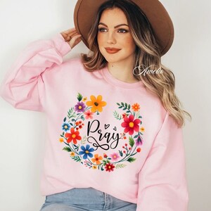 May include: Pink sweatshirt with a floral wreath design and the word "Pray" in black lettering.