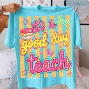 May include: A light blue t-shirt with the text "It's a good day to teach" in a colorful design. The design includes a stack of books, stars, and a bow. The shirt has orange and light blue vertical stripes.