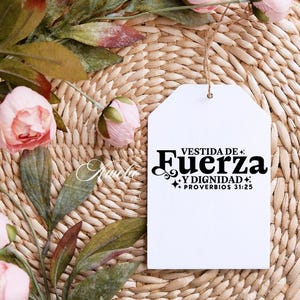 May include: A white tag with the text "VESTIDA DE FUERZA Y DIGNIDAD + PROVERBIOS 31:25" in black script. The tag hangs from a natural twine string. The background is a woven wicker mat, with pink flowers and green leaves.
