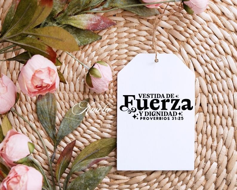 May include: A white tag with the text "VESTIDA DE FUERZA Y DIGNIDAD + PROVERBIOS 31:25" in black script. The tag hangs from a natural twine string. The background is a woven wicker mat, with pink flowers and green leaves.