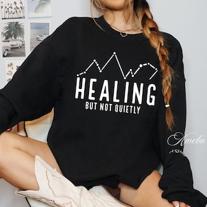 Puede incluir: Sudadera negra con texto blanco que dice "HEALING BUT NOT QUIETLY" y un gr&aacute;fico de una cordillera con estrellas.