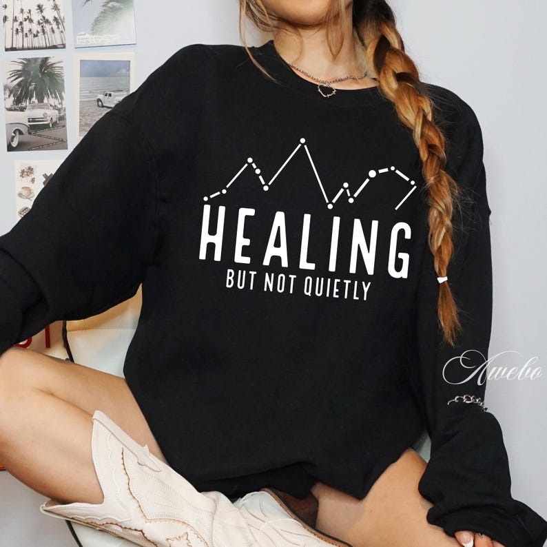 Puede incluir: Sudadera negra con texto blanco que dice "HEALING BUT NOT QUIETLY" y un gr&aacute;fico de una cordillera con estrellas.