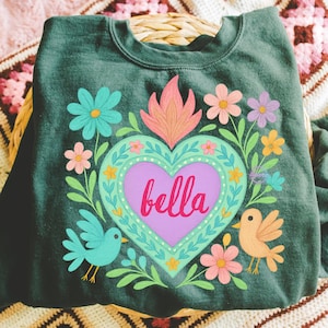 May include: Dark green sweatshirt featuring a heart design with the name "bella" in pink script. The heart is surrounded by colorful flowers, leaves, and two birds. The design has a folk art style.