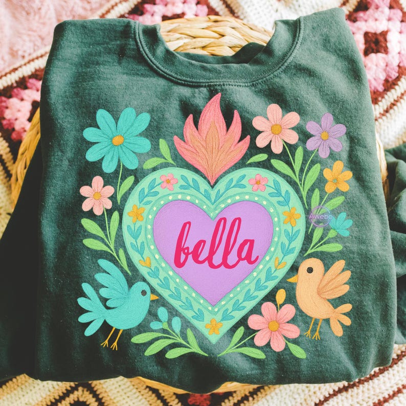 May include: Dark green sweatshirt featuring a heart design with the name "bella" in pink script. The heart is surrounded by colorful flowers, leaves, and two birds. The design has a folk art style.