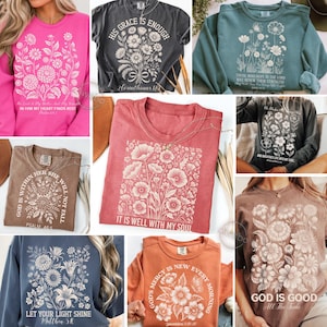 May include: A collection of various-colored t-shirts and sweatshirts, each featuring floral designs and inspirational text. The shirts are in shades of pink, gray, teal, red, brown, and orange. The text includes phrases like "His Grace is Enough" and "It is Well With My Soul."