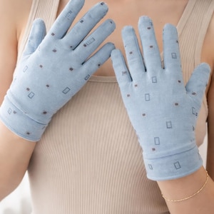 May include: Light blue gloves with a repeating pattern of small brown squares and rectangles. The gloves are made of a soft fabric and have a fitted cuff. The gloves are worn by a person wearing a beige tank top.