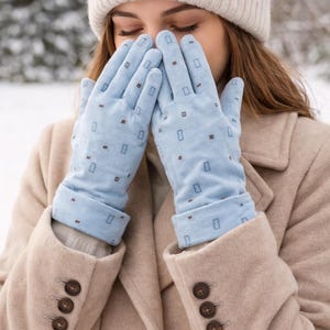 Può includere: Guanti azzurri con un motivo ripetuto di piccoli rettangoli marroni. I guanti sono indossati con un cappotto beige e un berretto di maglia beige chiaro. Il cappotto ha tre bottoni marroni sulla manica.