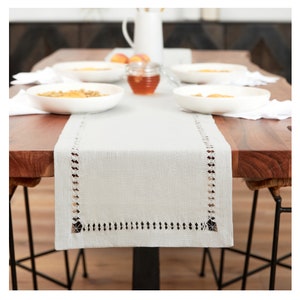May include: A light gray linen table runner with a delicate white embroidered border. The runner is laid on a wooden table with white placemats and bowls.