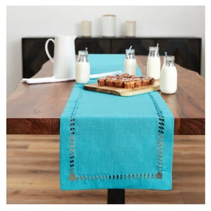 May include: A turquoise table runner with a decorative scalloped edge. The runner is laid on a wooden table with a pitcher, milk bottles, and a plate of pastries.
