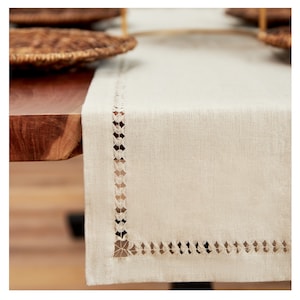 May include: A beige linen table runner with a delicate white lace trim. The runner is on a wooden table.