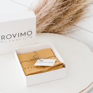 以下が含まれることがあります： 上部に「PROVIMO HOME」という文字が書かれた白い箱。箱が開いており、中には紐で結ばれた折り畳まれたマスタードイエローのリネンキッチンクロスと、「PROVIMO」という文字が書かれた小さな白いタグが入っています。