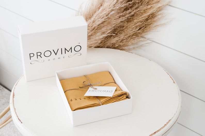 以下が含まれることがあります： 上部に「PROVIMO HOME」という文字が書かれた白い箱。箱が開いており、中には紐で結ばれた折り畳まれたマスタードイエローのリネンキッチンクロスと、「PROVIMO」という文字が書かれた小さな白いタグが入っています。