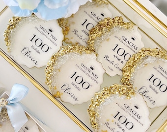 May include: Round white cookies with gold leaf edges, featuring the text 100 Birthday and names like Olivier or Mamita Carmen. Some are in individual boxes with light blue ribbons, while others are displayed in a gold-trimmed glass tray.