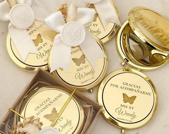 May include: A collection of gold-colored compact mirrors with butterfly designs and the text "Gracias Por Acompañarme" and "Mis XV Wendy" on a white tray. Each mirror is adorned with a white ribbon and a wax seal. The tray is surrounded by decorative elements.