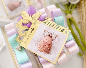 Puede incluir: Una pequeña caja transparente llena de cintas de colores pastel y una placa dorada con el nombre "Amaris" y la fecha "Nov 16, 2021". La placa también presenta una pequeña cruz y un diseño de mariposa.
