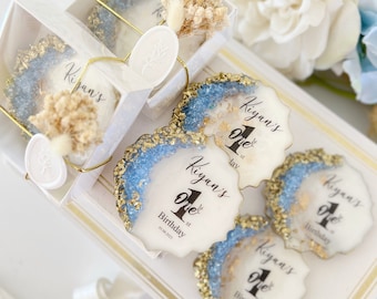 May include: Round white resin coasters with scalloped edges, blue and gold glitter accents, and black text reading Kiyan's 1st Birthday. Some are packaged in clear boxes with gold string, white wax seals, and dried flowers, while others are loose.