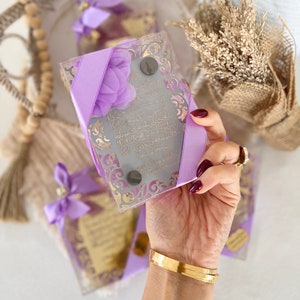 May include: A set of four decorative glass boxes with purple ribbon and a floral design. Each box contains a small, framed piece of paper with Arabic calligraphy. The boxes are arranged on a woven surface.