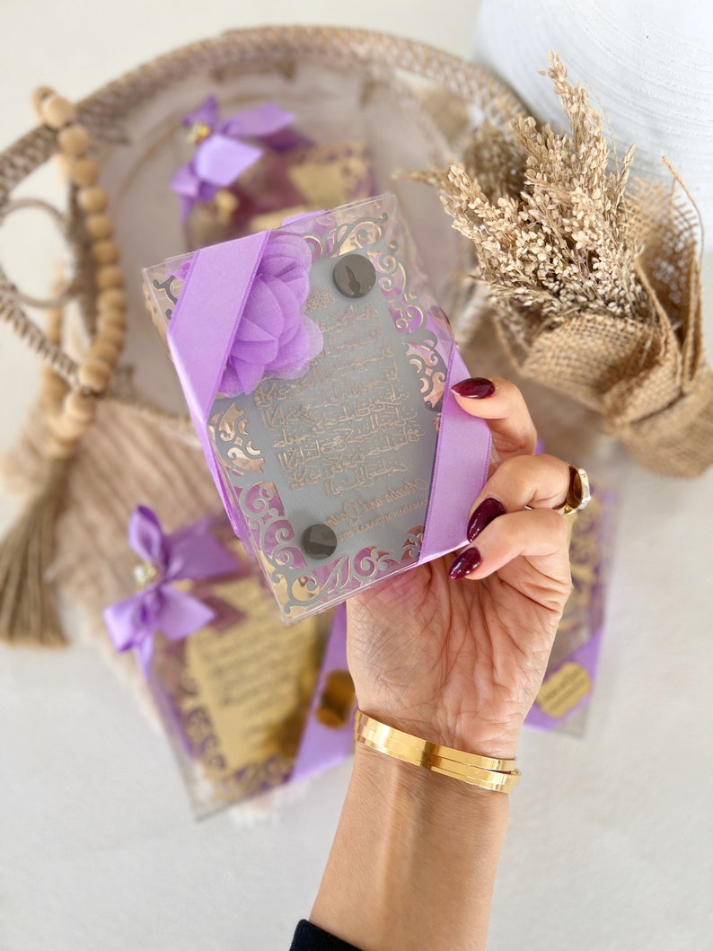 May include: A set of four decorative glass boxes with purple ribbon and a floral design. Each box contains a small, framed piece of paper with Arabic calligraphy. The boxes are arranged on a woven surface.