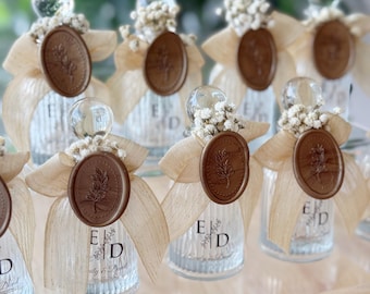 Puede incluir: Botellas de vidrio transparente con un diseño decorativo, coronadas con una bola de vidrio transparente y pequeñas flores blancas. Cada botella tiene un sello de cera marrón con un diseño floral y una cinta. Las botellas tienen las letras "E" y "D".