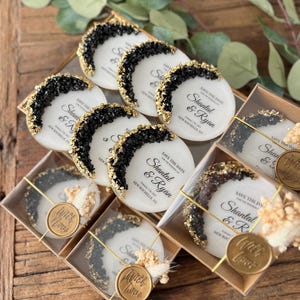 May include: Save the date wedding favors with black and gold glitter accents. The favors are round and feature the text "Save the Date" and the names "Shantal & Ryan" with the date and location. The favors are packaged in clear boxes with a gold wax seal that says "With Love".