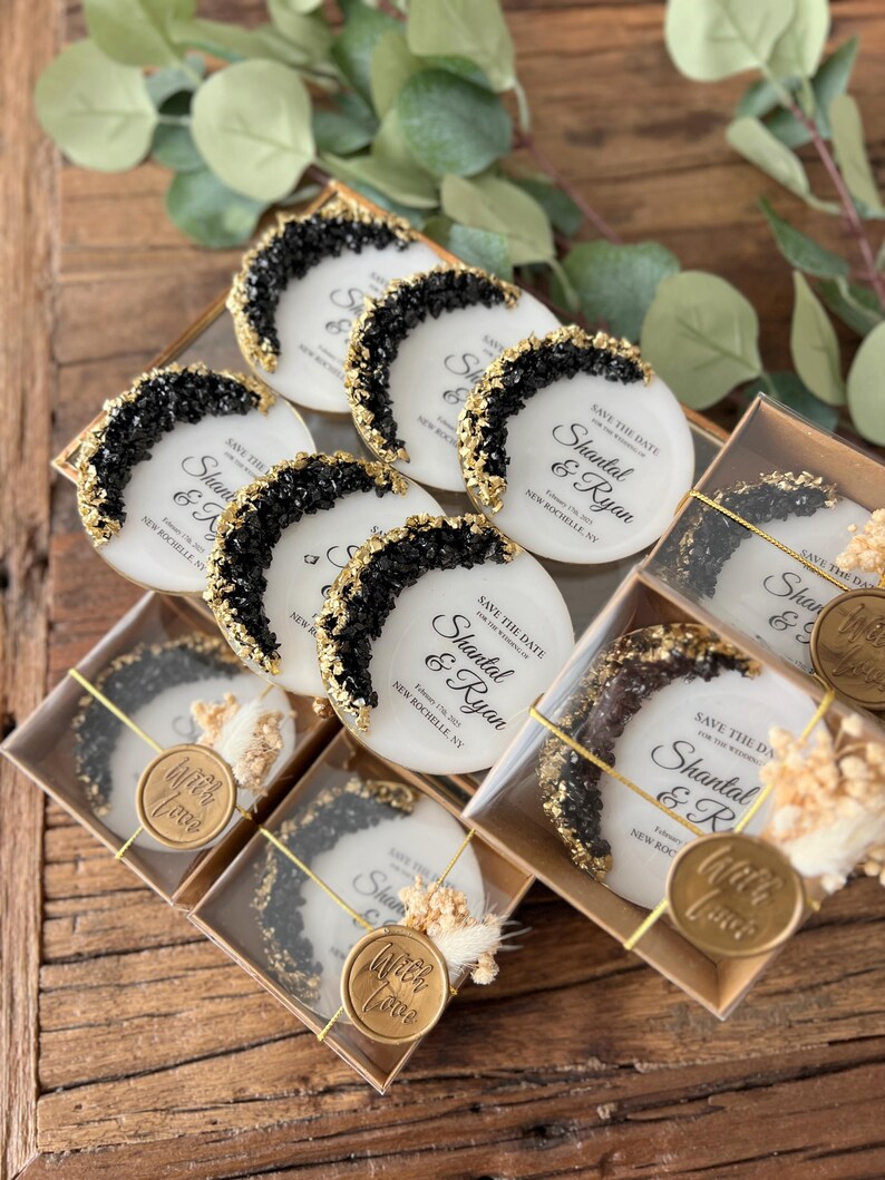 May include: Save the date wedding favors with black and gold glitter accents. The favors are round and feature the text "Save the Date" and the names "Shantal & Ryan" with the date and location. The favors are packaged in clear boxes with a gold wax seal that says "With Love".