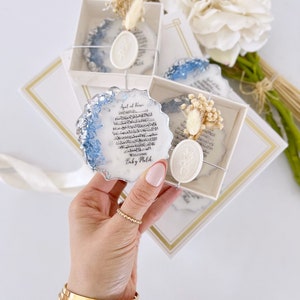 May include: A white box with a blue and silver resin coaster featuring Arabic calligraphy and a wax seal. The coaster is decorated with dried flowers and the text "Welcome Baby Malik".